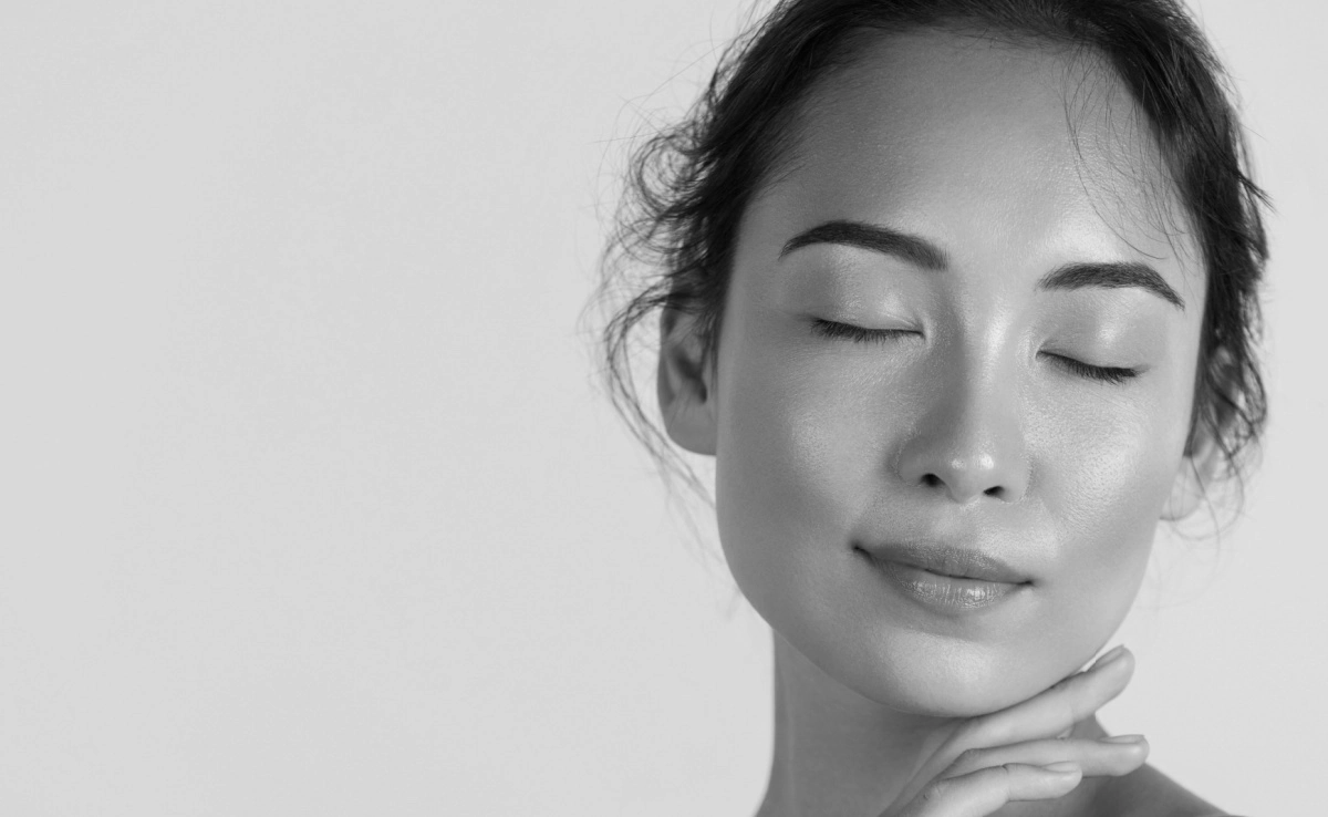 Beautiful asian girl model touching fresh glowing hydrated facial skin on beige background closeup.